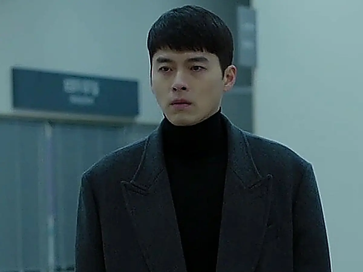 A man in a dark tailored coat and black turtleneck standing motionless in a brightly lit hallway